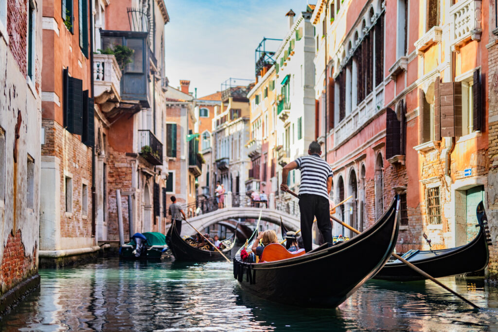 Gondel mit Gondoliere fährt durch einen engen Kanal in Venedig, flankiert von terrakottafarbenen Palazzi und einer kleinen Bogenbrücke.