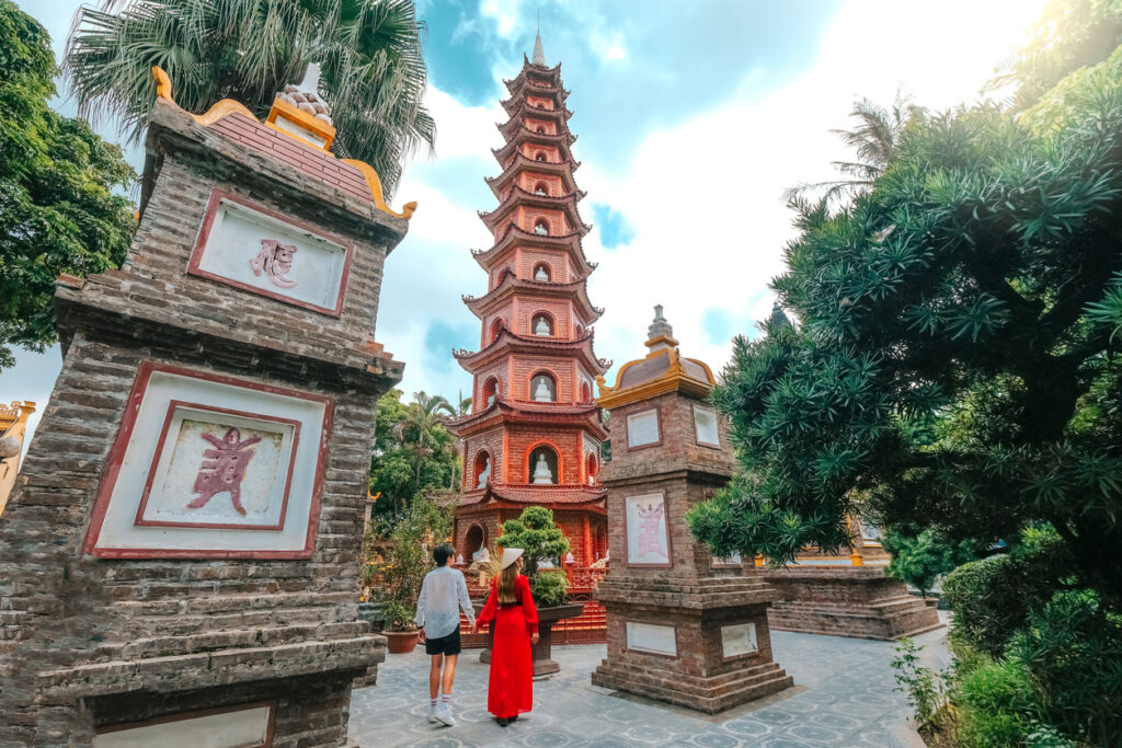 Rote, mehrstöckige Tran-Quoc-Pagode mit Ziegelstupas und zwei Besuchern im Hof, umgeben von üppigem Grün und blauem Himmel.
