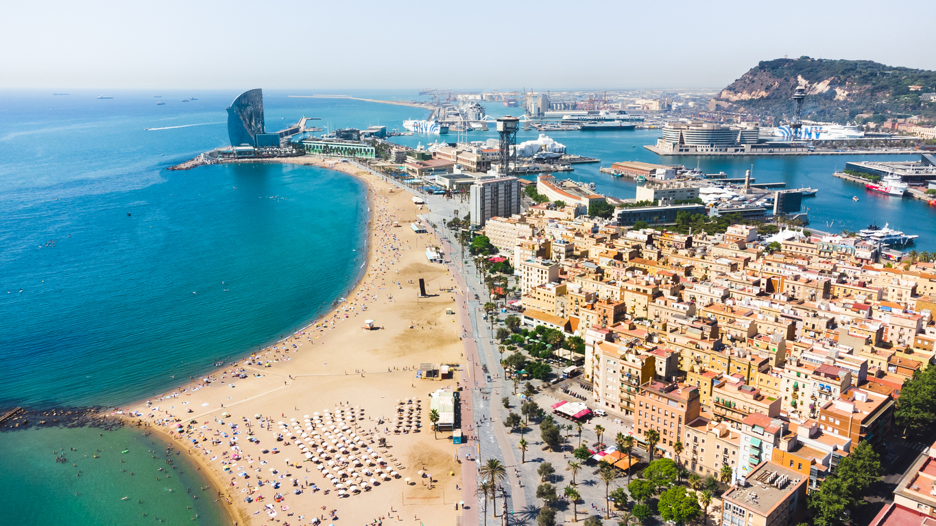 Luftaufnahme der Bucht von Barceloneta: goldener Sandstrand mit Sonnenschirmen und Badenden, rechts das Stadtviertel Barceloneta, im Hintergrund das segelförmige W-Hotel, Hafenbecken von Port Vell und der Hügel Montjuïc.