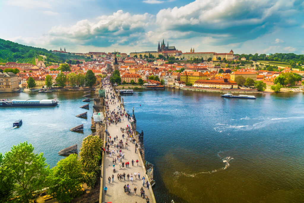 Luftblick auf die Karlsbrücke mit vielen Fußgängern, Booten auf der Moldau und der Prager Burg im Hintergrund.