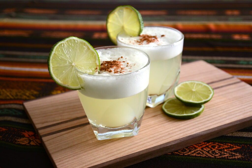 Zwei Gläser Pisco Sour mit Limettenscheiben, heller Schaumschicht und brauner Garnitur auf einem Holzbrett.