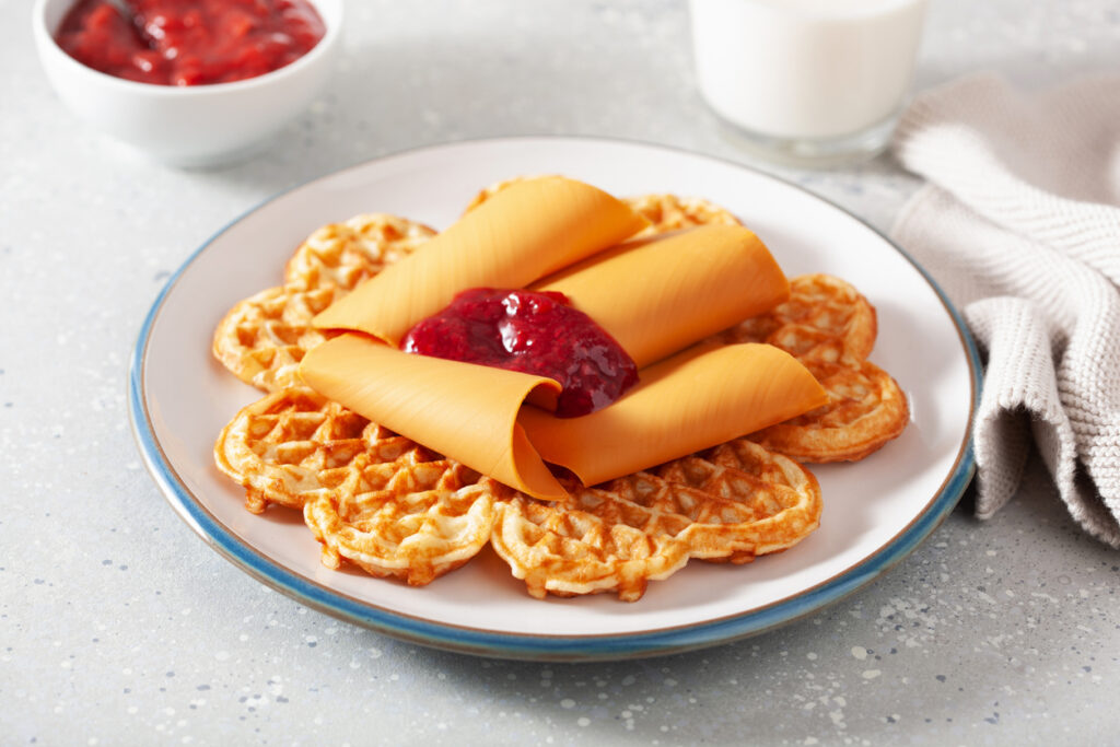 Auf einem Teller liegen Herzwaffeln, belegt mit drei gerollten Scheiben norwegischem Brunost und einem Klecks Himbeermarmelade.