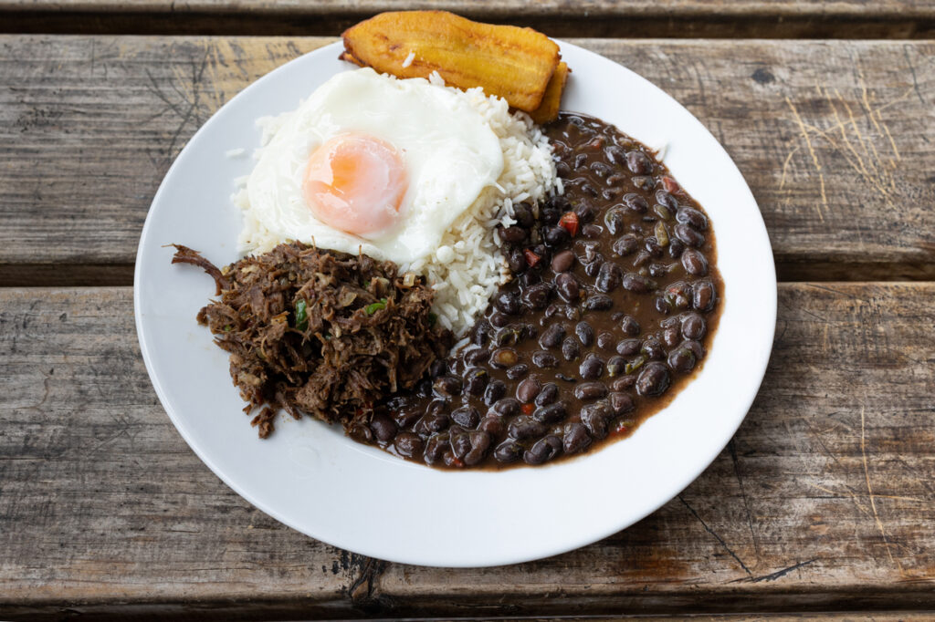 Weißer Teller mit Pabellón criollo: Reis mit Spiegelei, geschmortem Rindfleisch, schwarzen Bohnen und gebratener Kochbanane.