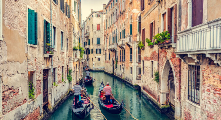 Gondolieri steuern drei Gondeln durch einen schmalen Kanal in Venedig, flankiert von verwitterten Backsteinfassaden mit Balkonen und Fensterläden.