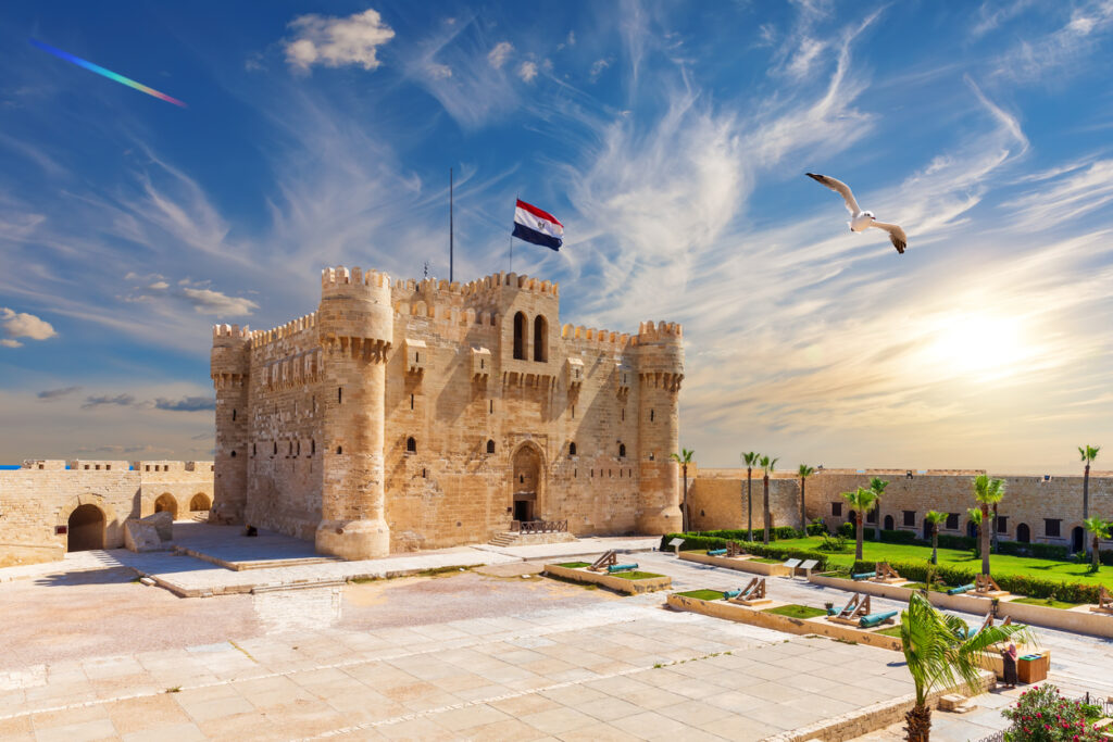 Die mittelalterliche Festung Qaitbay in Alexandria mit ägyptischer Flagge, Innenhof, Palmen und Möwe vor blauem Himmel und tief stehender Sonne.