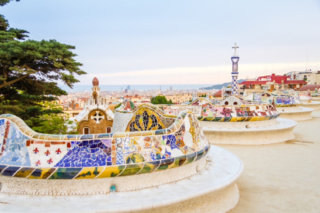 Geschwungene, trencadís-bedeckte Sitzbänke von Gaudí auf der Terrasse des Park Güell, dahinter die Stadt bis zum Meer.