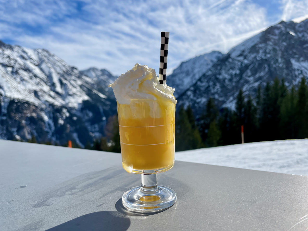Glas Bombardino mit Sahne und gestreiftem Strohhalm vor verschneiten Bergen.