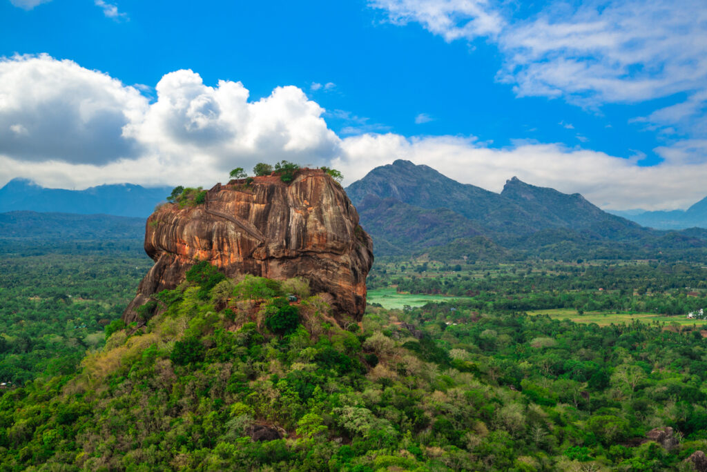 Luftaufnahme des Sigiriya-Löwenfelsens im Kulturdreieck Sri Lankas mit Terrassen, Treppen und umgebendem Dschungel unter blauem Himmel.