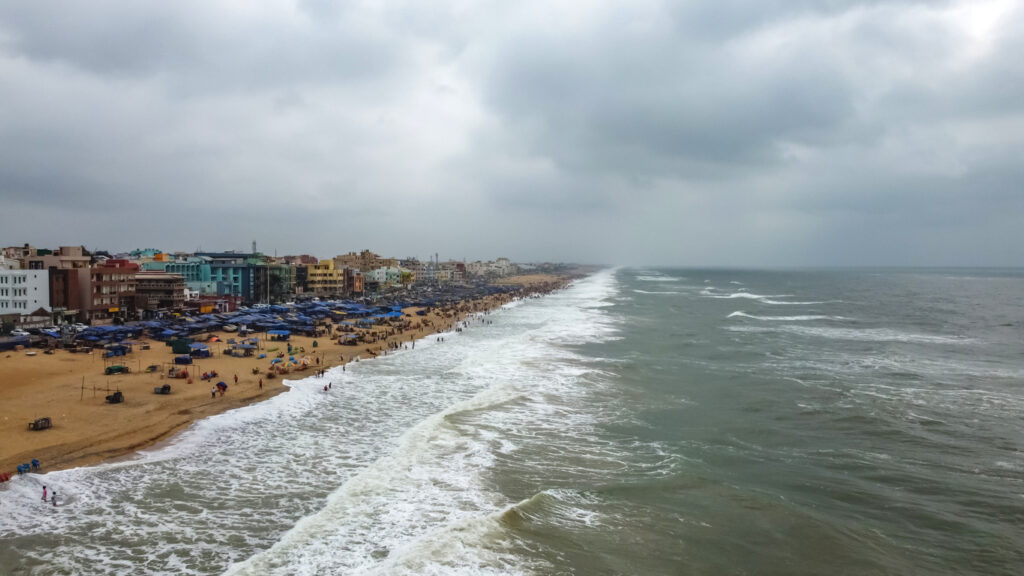 Blick auf den weitläufigen Strand von Puri mit Wellen, dicht stehenden Strandständen und bunten Häusern unter bedecktem Himmel.