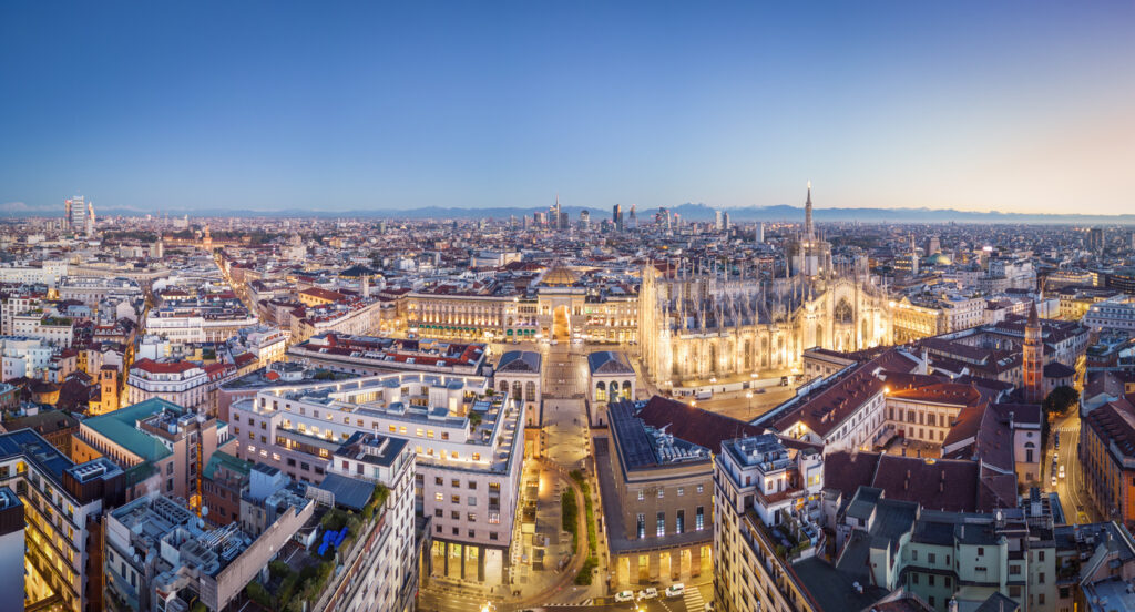 Panoramablick bei Abenddämmerung auf den erleuchteten Dom, die Galleria und die Skyline mit Alpen im Hintergrund.