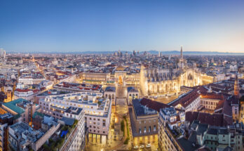 Panoramablick bei Abenddämmerung auf den erleuchteten Dom, die Galleria und die Skyline mit Alpen im Hintergrund.