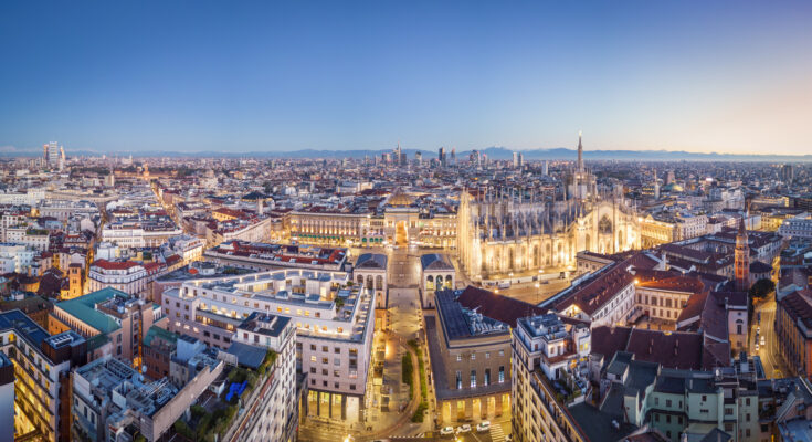 Panoramablick bei Abenddämmerung auf den erleuchteten Dom, die Galleria und die Skyline mit Alpen im Hintergrund.