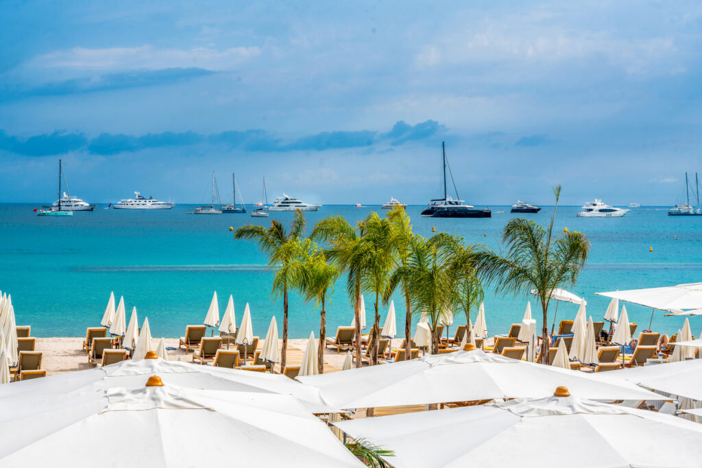 Panoramablick von der Promenade de la Croisette über weiße Sonnenschirme, Palmen und Liegen auf das türkisfarbene Mittelmeer mit Segel- und Motoryachten in der Bucht von Cannes.