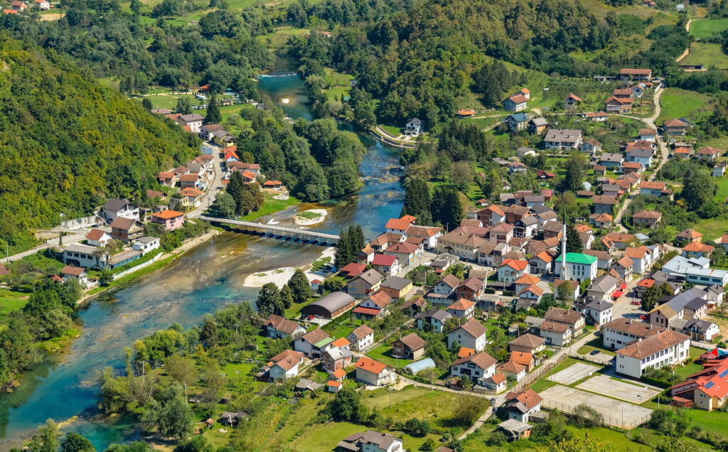 Luftaufnahme eines Ortes mit Minarett, Brücke und vielen Häusern entlang der türkisfarbenen Una inmitten grüner Hügel.