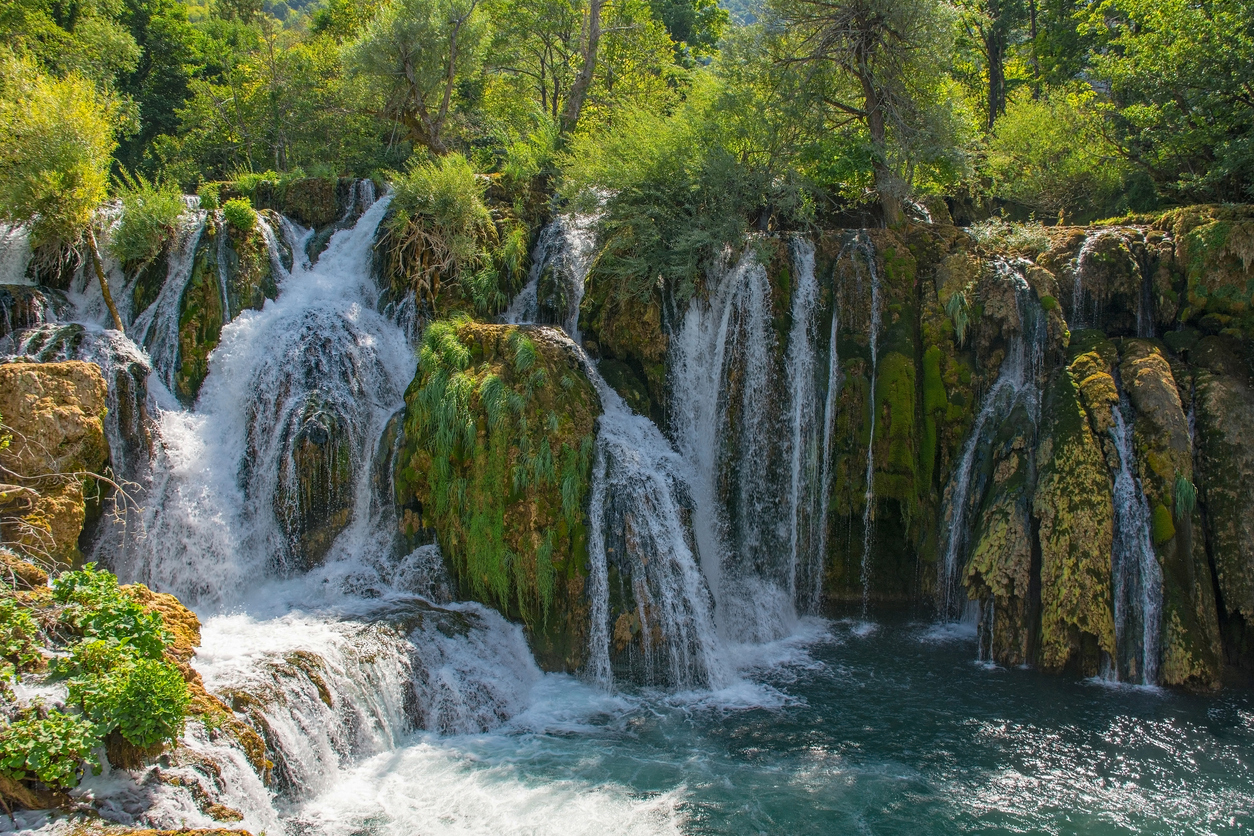 Mehrere Wasserfälle stürzen über moosbewachsene Felsen in ein türkisblaues Becken im Una-Nationalpark.