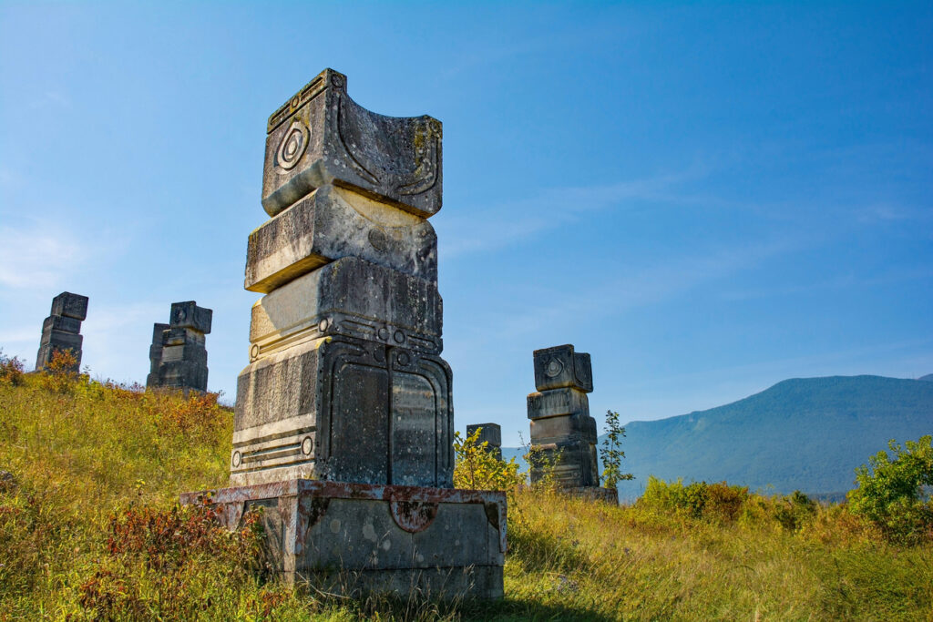 Mehrere verzierte Steinpfeiler stehen auf einer grasigen Anhöhe vor bewaldeten Bergen im Una-Nationalpark.