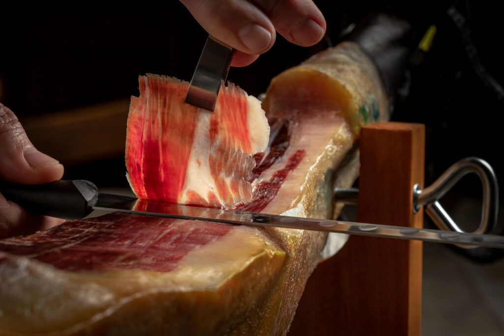 Nahaufnahme: hauchdünne Scheibe Jamón Ibérico wird mit langem Messer vom Schinkenbein geschnitten.