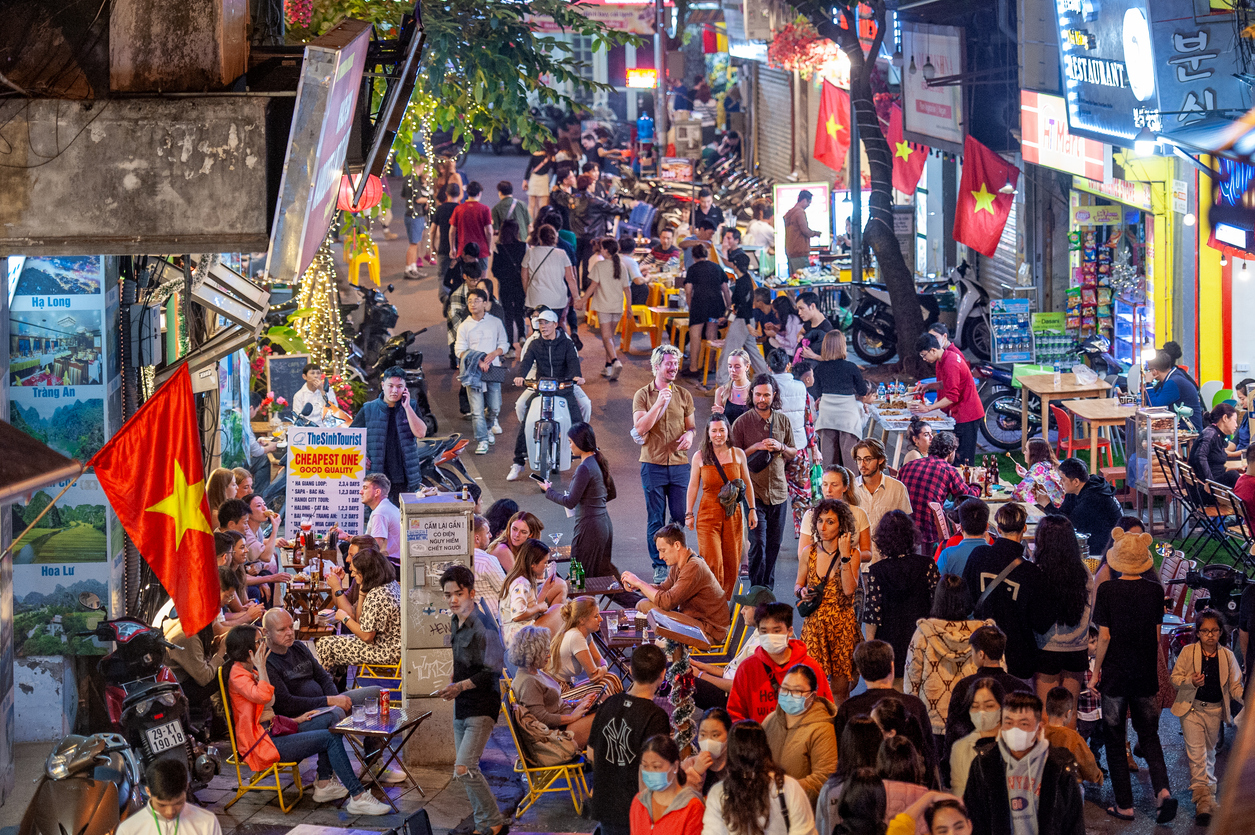 Belebte Gasse mit Streetfood-Ständen, vielen Menschen und vietnamesischen Fahnen am Abend.