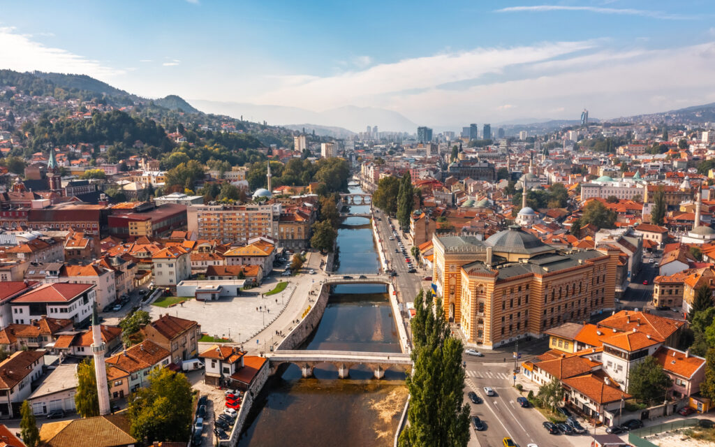 Panoramablick über Sarajevo mit der Miljacka in der Bildmitte, rechts der Vijećnica und roten Dächern vor grünen Hügeln.