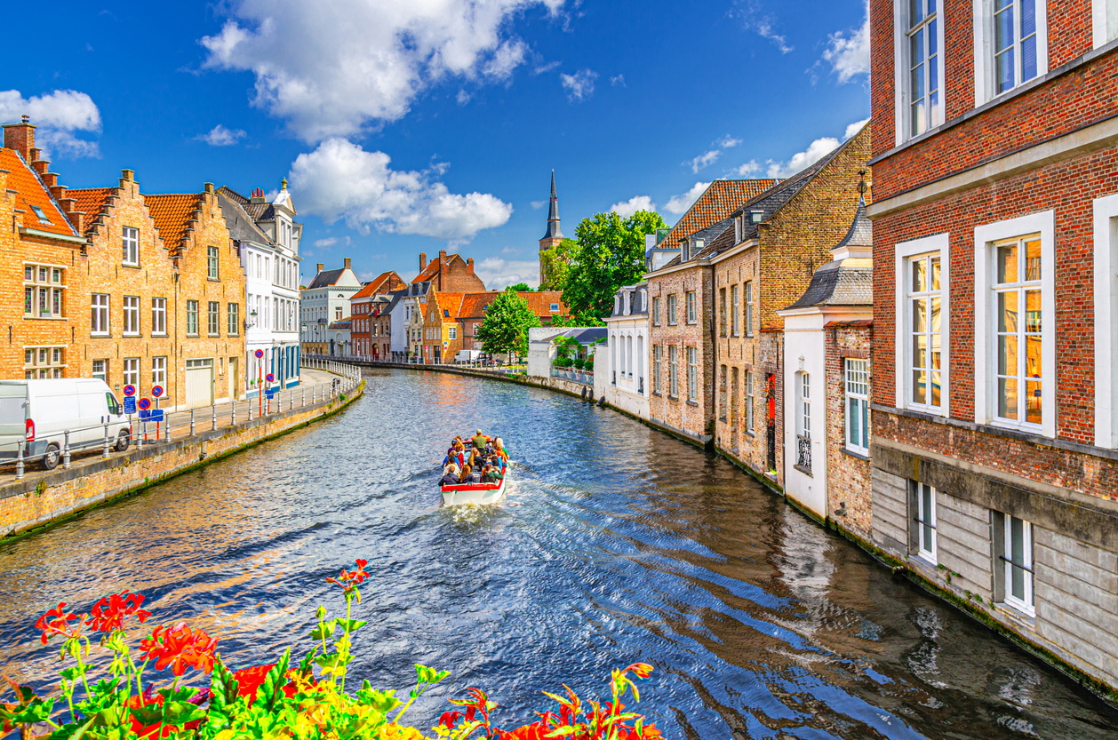 Kanal in Brügge mit Backsteinhäusern, Giebeln, Ausflugsboot und blauem Himmel mit Wolken.