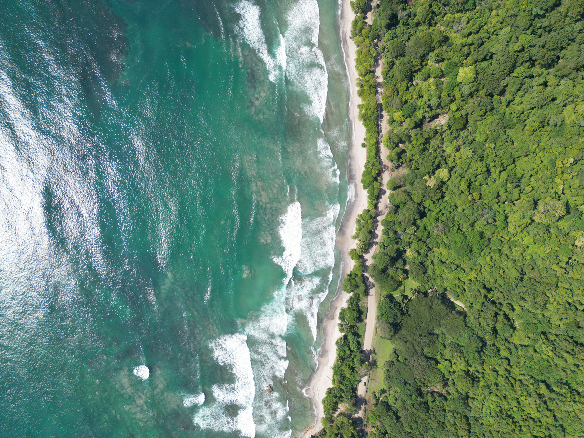 Luftaufnahme von Santa Teresa: dichter Wald rechts, heller Sandstreifen und türkisgrünes Meer mit brechenden Wellen links.