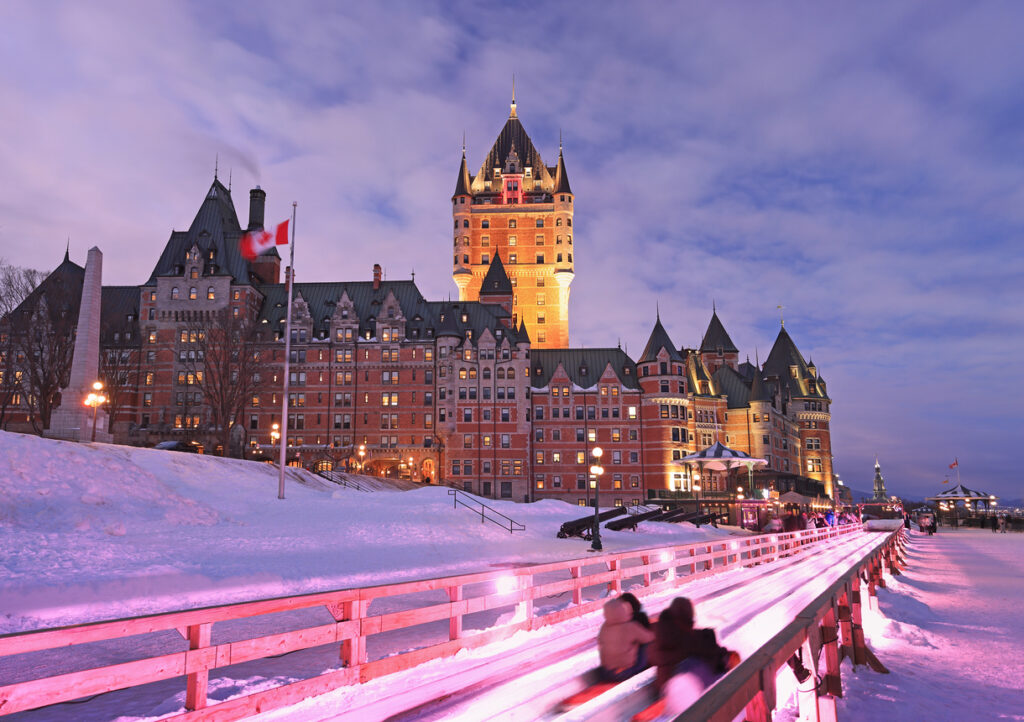Vor dem beleuchteten Château Frontenac sausen Menschen auf der Holzrodelbahn durch den Schnee.