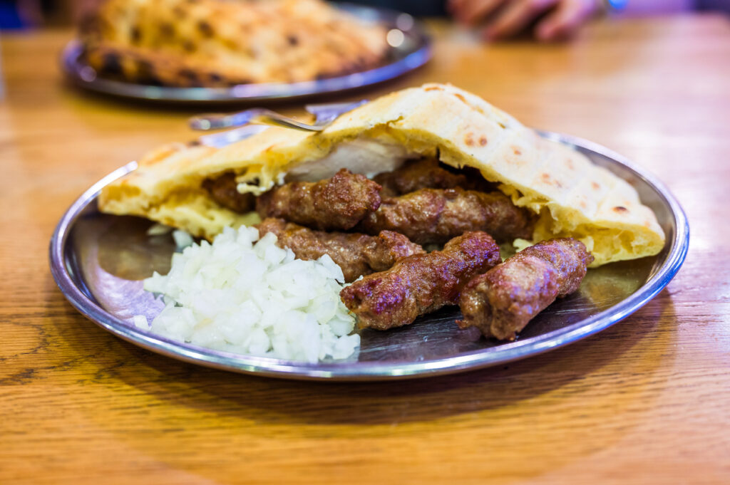 Ćevapi im aufgeschlagenen Fladenbrot mit gehackten Zwiebeln auf einem Metallteller, aufgenommen in einem Sarajevaner Lokal.