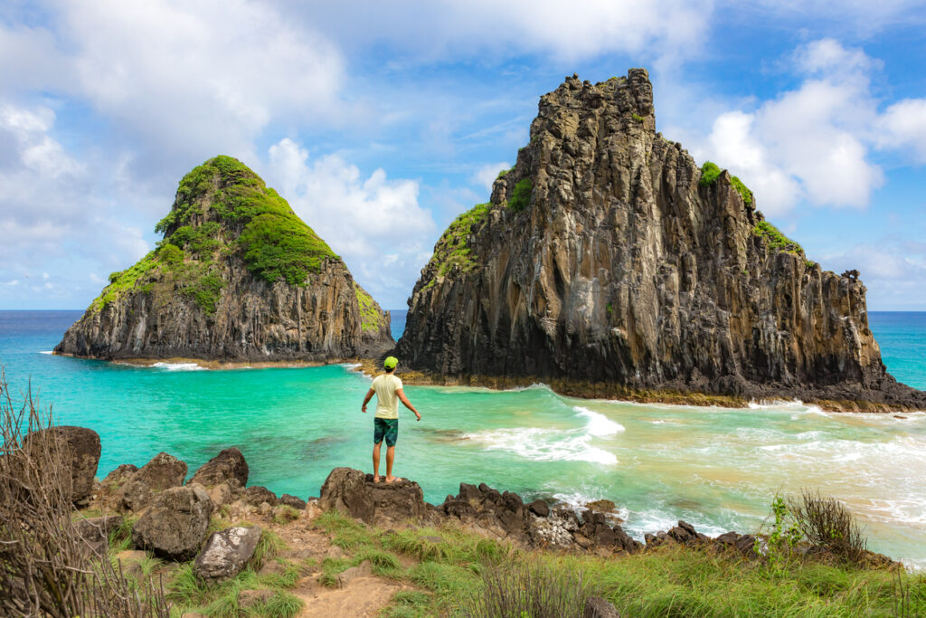 Person steht auf Felsen und blickt auf türkisgrüne Bucht mit den Morro-Dois-Irmãos-Felsen auf Fernando de Noronha.