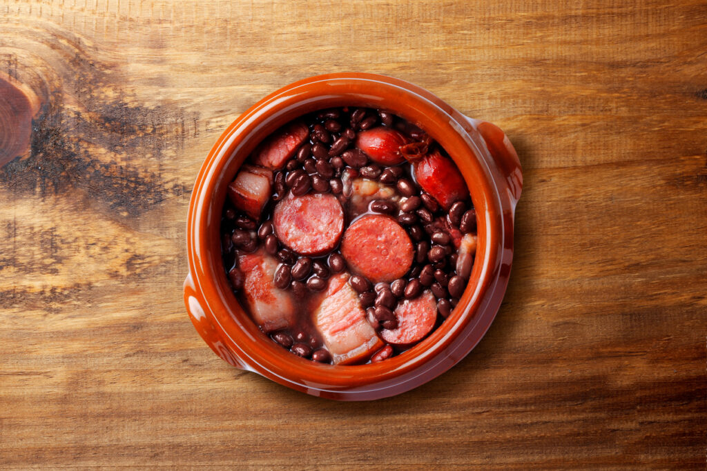 Tontopf mit Feijoada: schwarze Bohnen, Wurstscheiben und Fleischstücke auf Holzuntergrund.