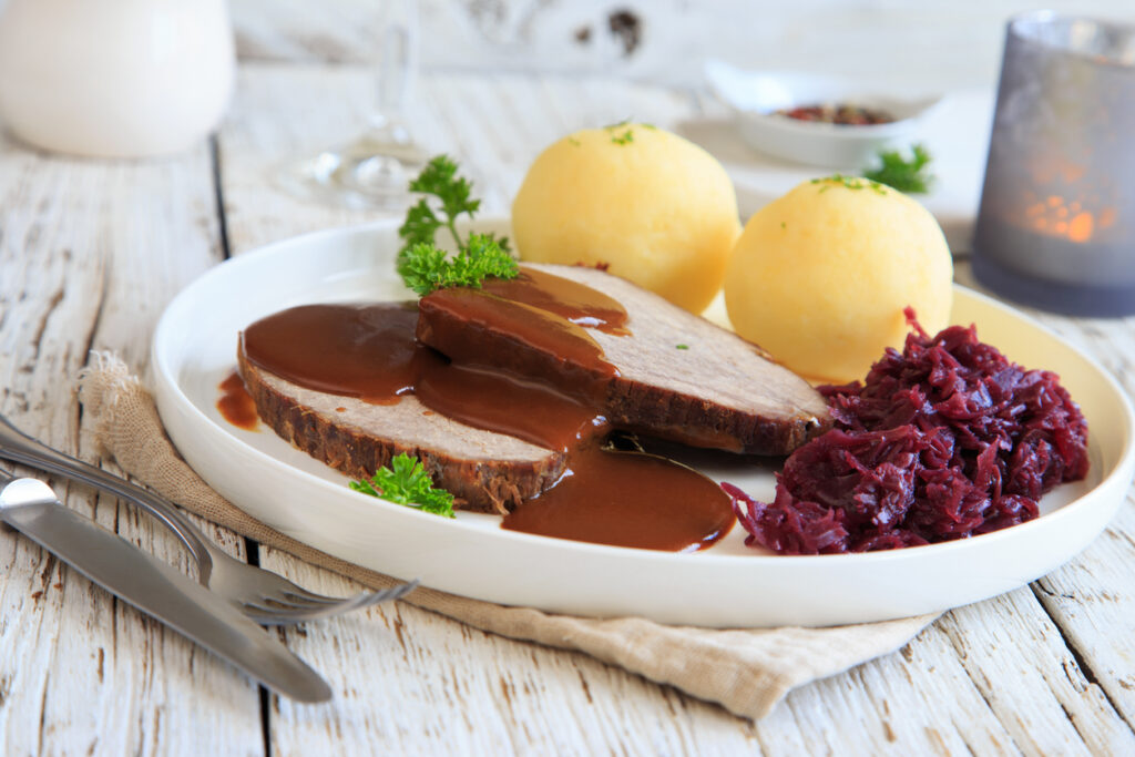 Teller mit aufgeschnittenem Sauerbraten, dunkler Sauce, Kartoffelklößen und Rotkohl.