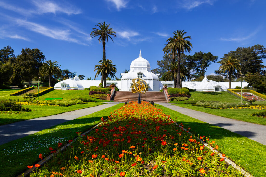 Das weiße viktorianische Conservatory of Flowers im Golden Gate Park, flankiert von Palmen, Rasenflächen und bunten Blumenrabatten unter blauem Himmel.
