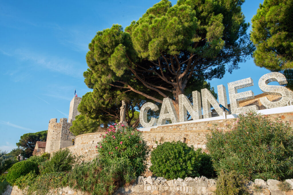 Großer CANNES-Schriftzug vor Pinien am Hügel Le Suquet mit mittelalterlichem Turm und französischer Flagge über Steinmauern unter blauem Himmel.