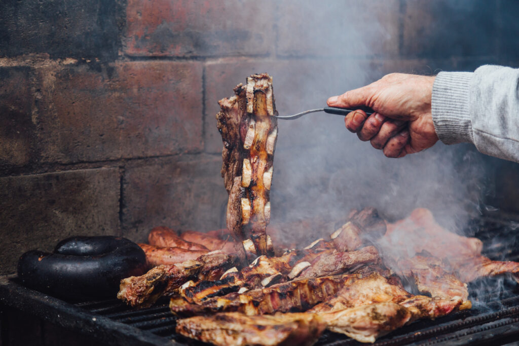 Eine Hand hebt ein Rinderrippenstück von der rauchigen Parrilla, daneben grillen Würste und weitere Fleischstücke.