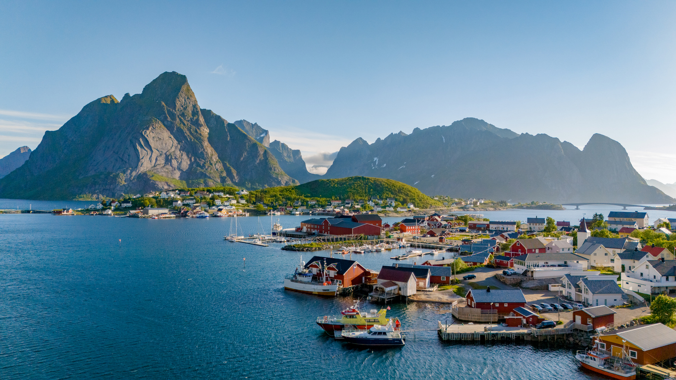 Luftaufnahme eines Lofoten-Hafens mit roten Rorbuer, Booten, Brücke und fjordartigen Wasserflächen vor schroffen Bergen.