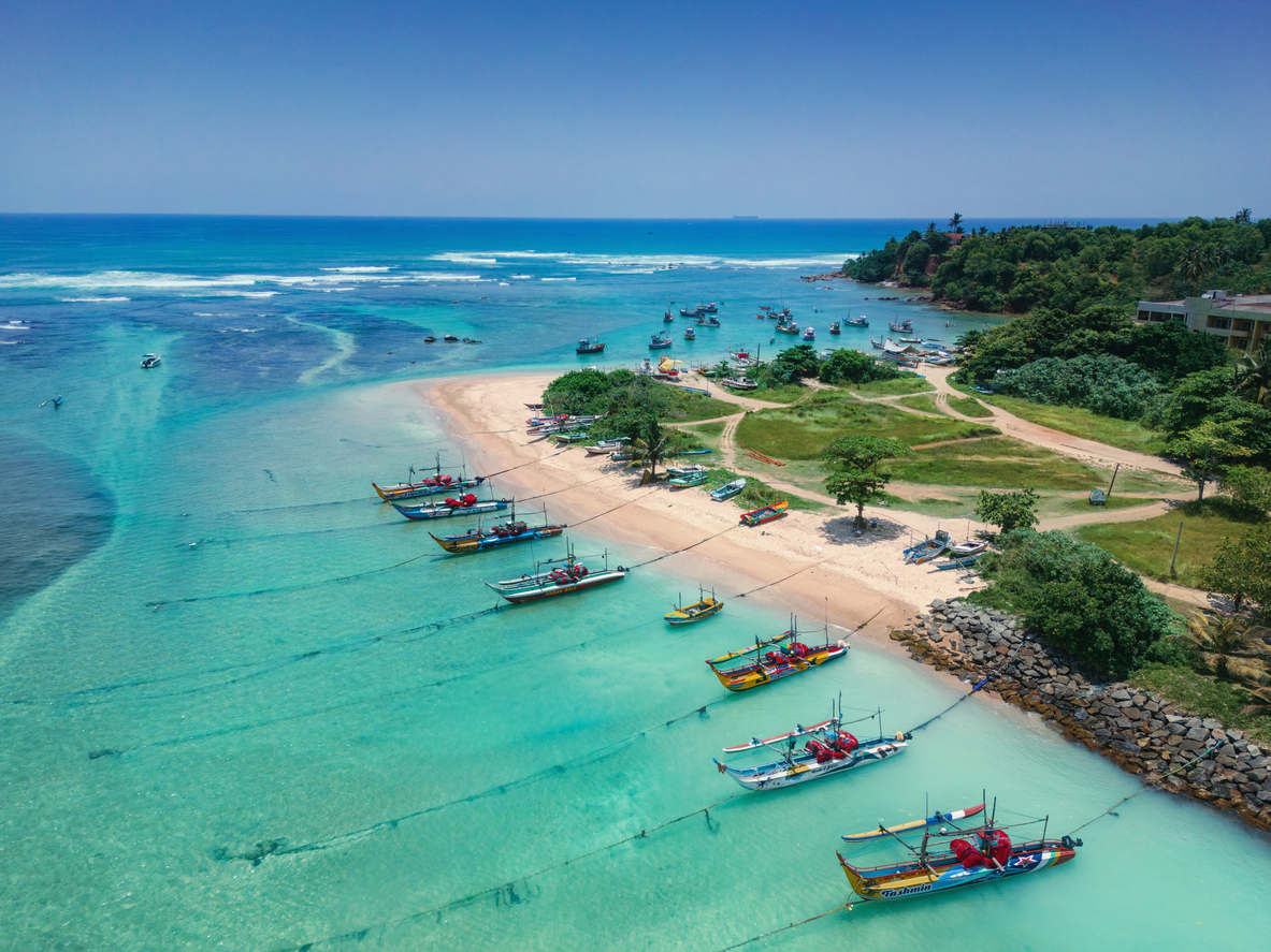 Luftaufnahme von Weligama mit sandiger Landzunge, klarem türkisblauem Wasser und zahlreichen farbigen Auslegerfischerbooten.