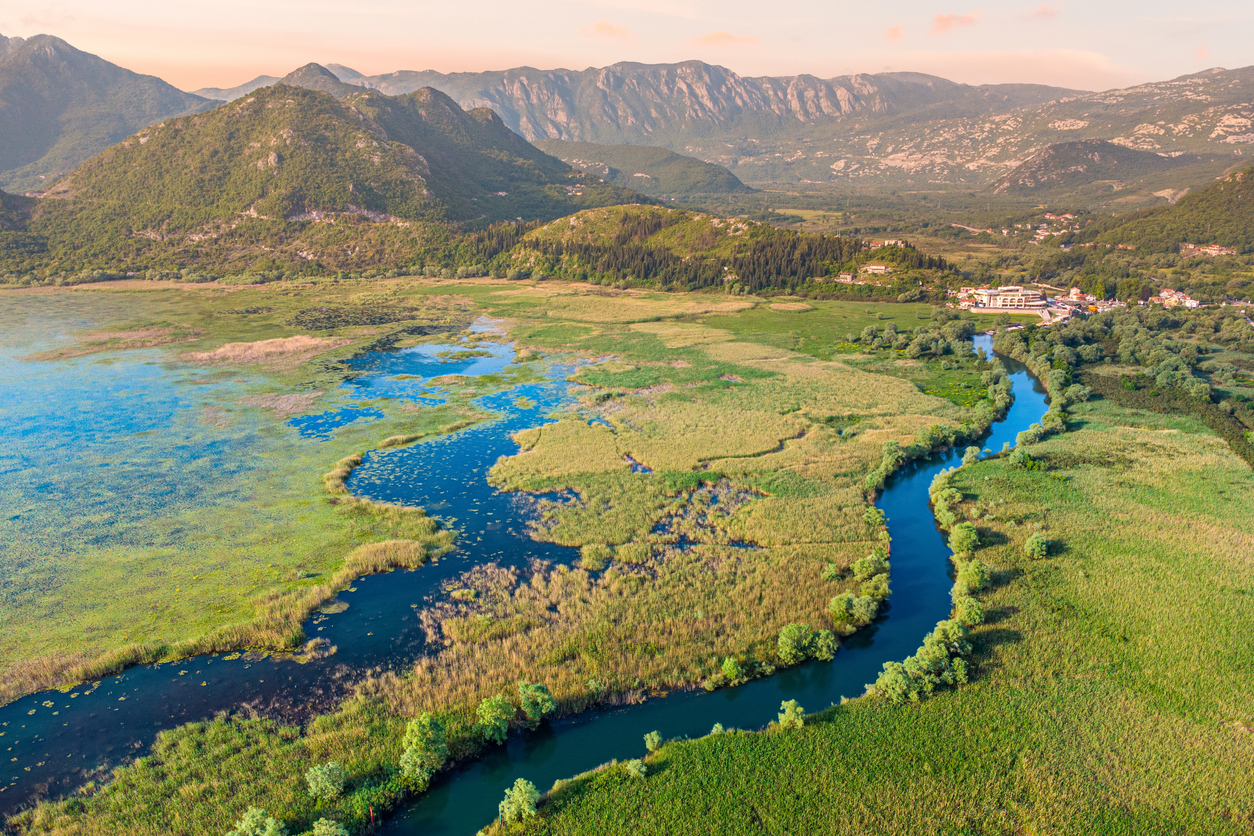 Luftaufnahme von Virpazar mit mäanderndem Kanal, weiten Schilfflächen, Seerosenteppichen und Bergkulisse im warmen Abendlicht.