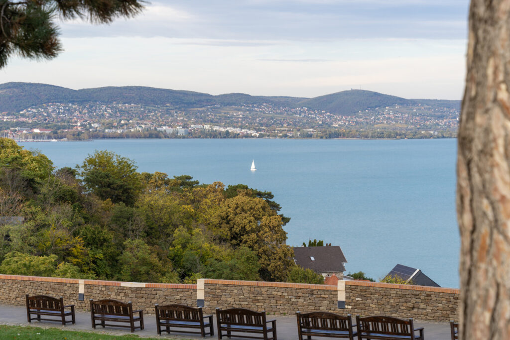 Aussichtspunkt in Balatonfüred mit Steinmauer und Sitzbänken im Vordergrund; darunter Baumkronen, dahinter der türkisfarbene Balaton mit einem weißen Segelboot und sanften Hügeln mit Ortschaften am gegenüberliegenden Ufer.