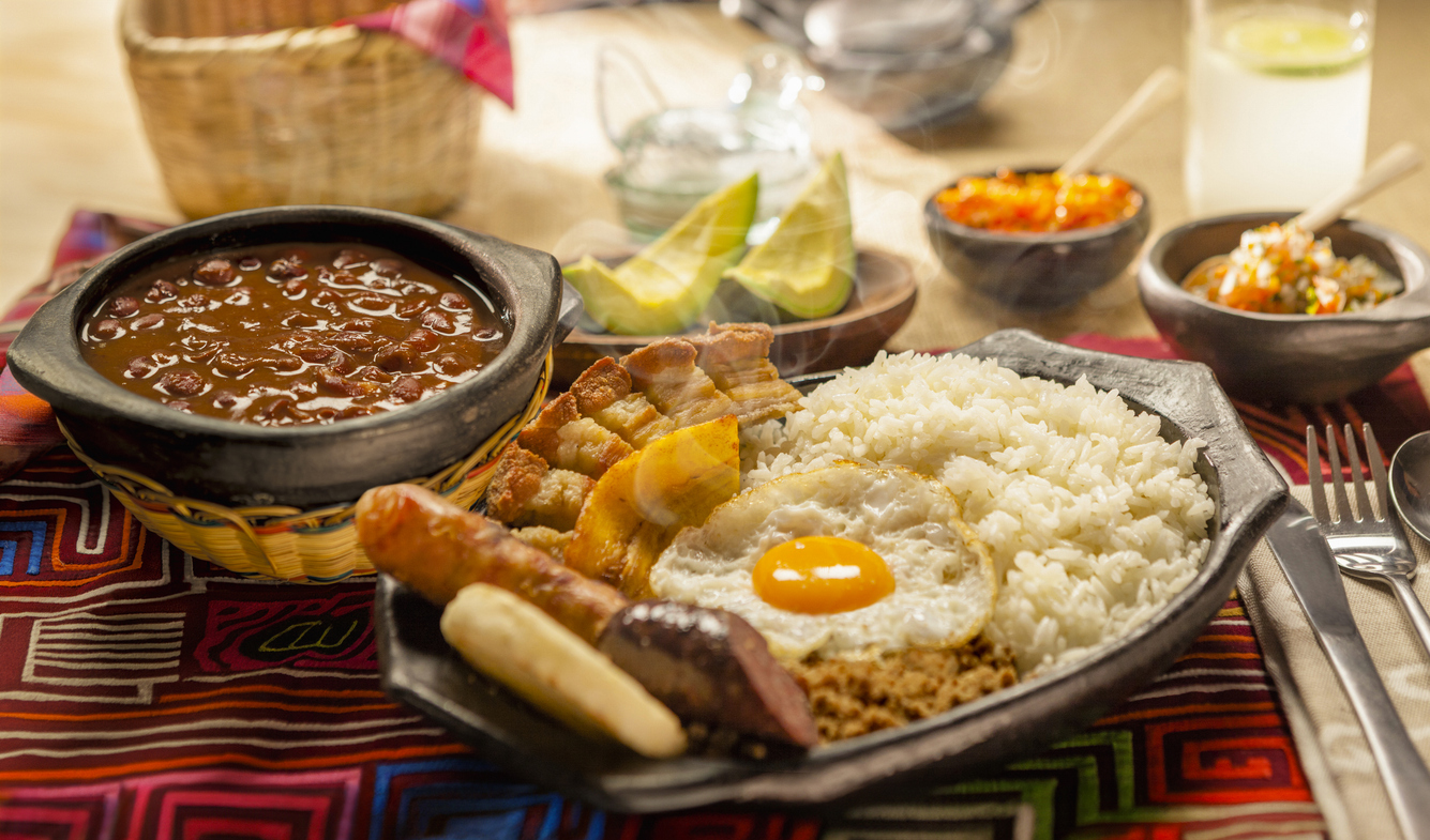 Teller mit Bandeja paisa: Reis, Spiegelei, Chicharrón, Chorizo, Kochbanane und Hack, daneben dampfende Bohnen.
