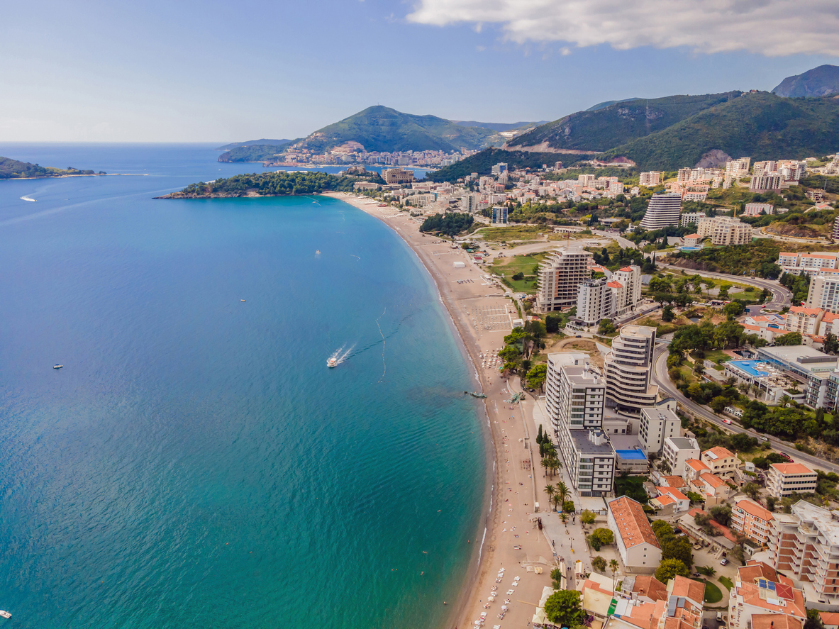 Luftaufnahme von Budva in Montenegro mit geschwungener Bucht, türkisblauem Meer, Promenade, Hotels und grünen Bergen im Hintergrund.