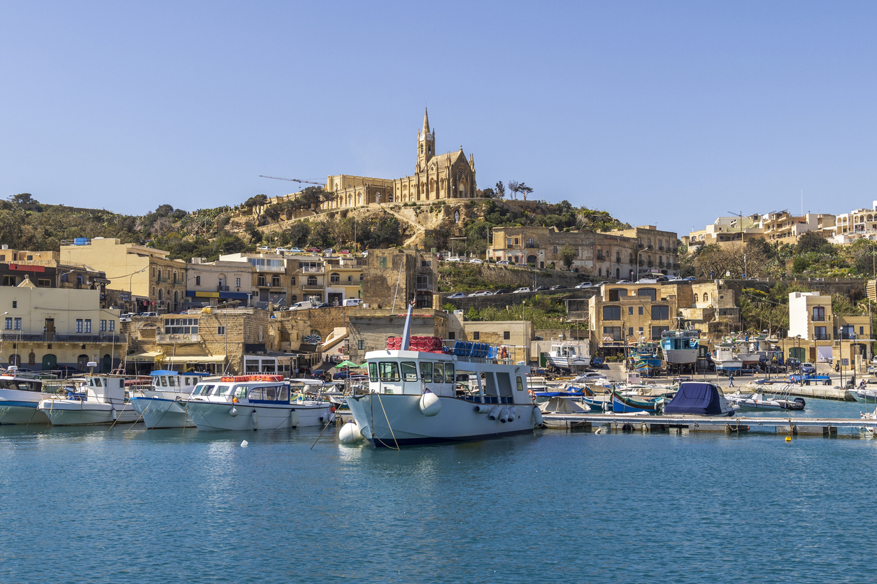 Blick auf den Hafen Mġarr auf Gozo mit bunten Booten, Kalksteinfassaden und der Kirche Unserer Lieben Frau von Lourdes auf dem Hügel.