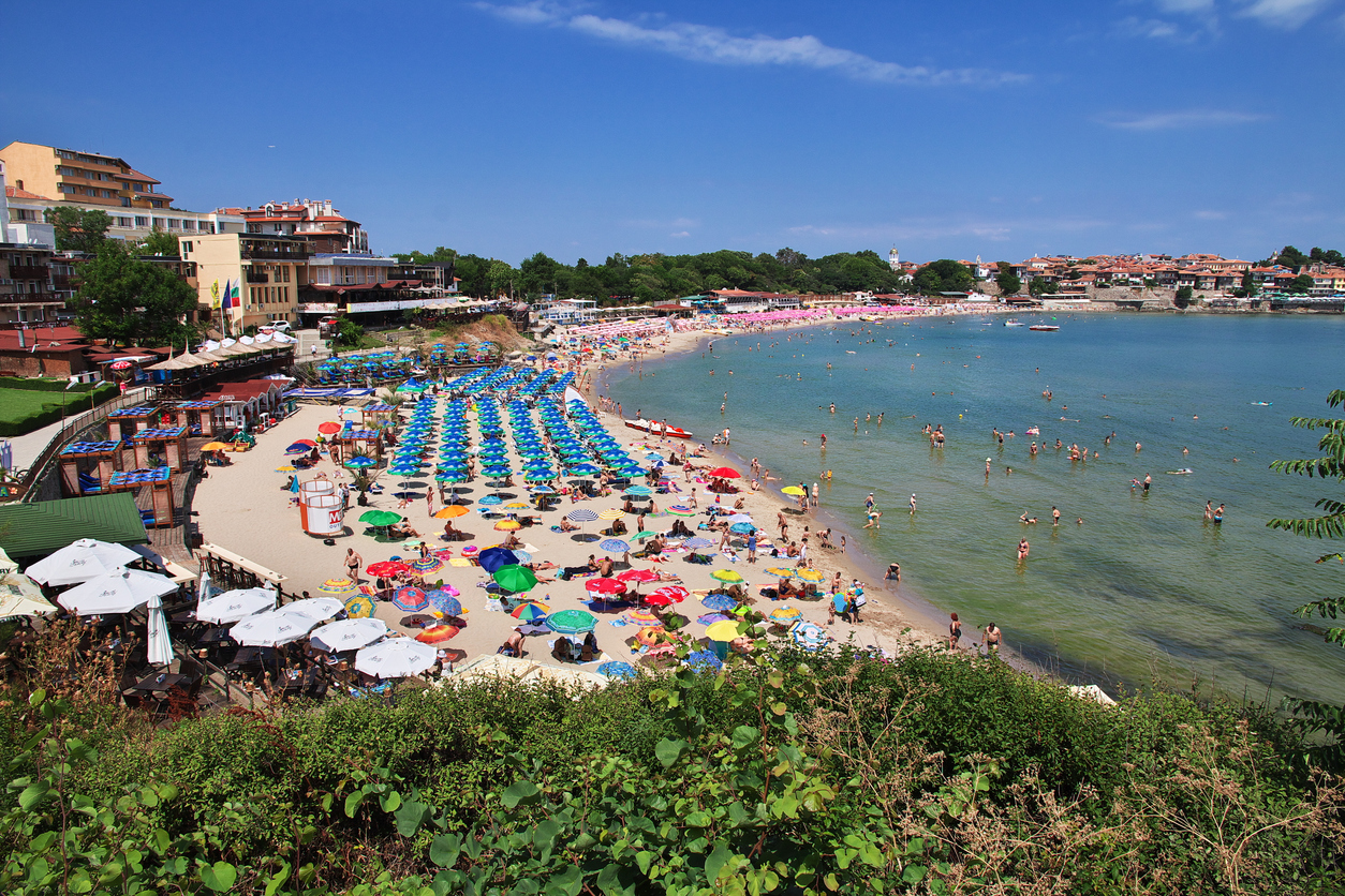 Bunter, belebter Strand mit vielen Sonnenschirmen, flachem türkisfarbenem Wasser und der Altstadt von Sozopol im Hintergrund.