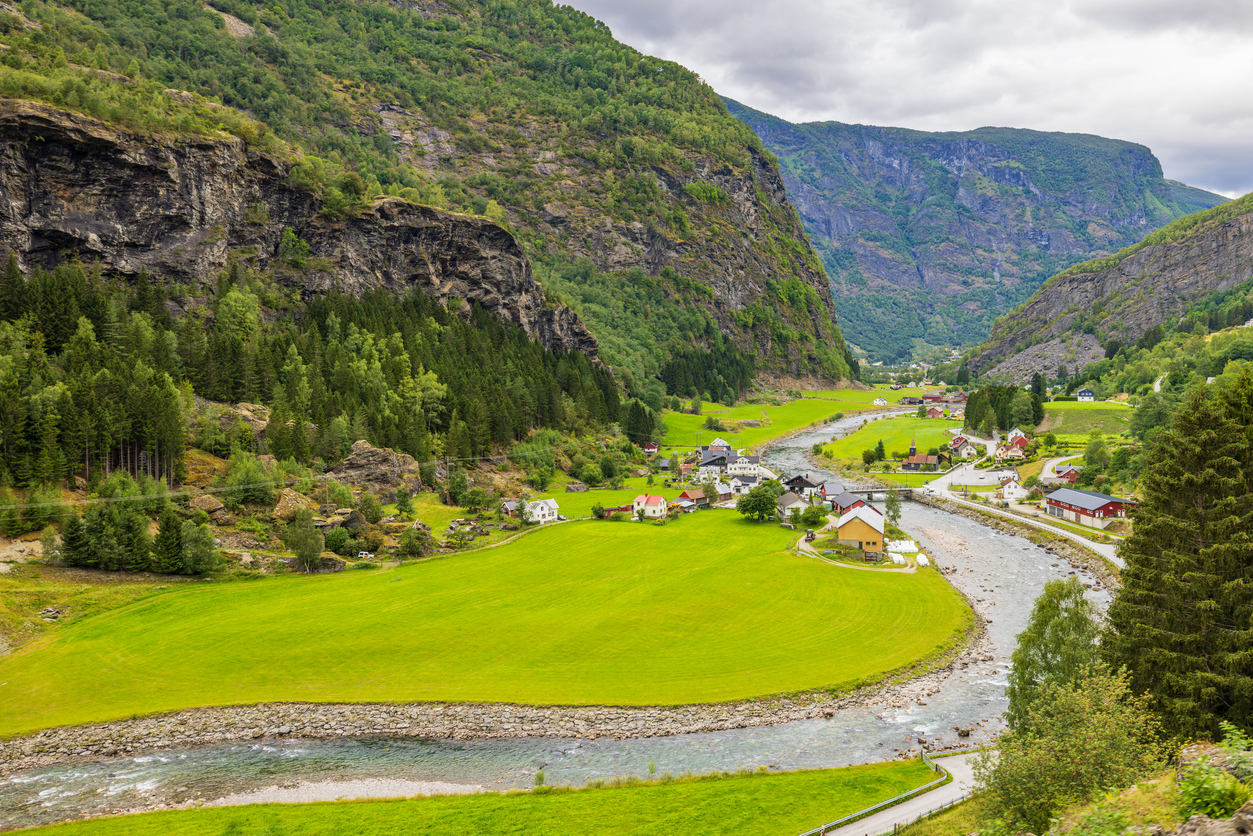 Weites Talpanorama von Flåm mit der sich windenden Flåmselvi, grünen Wiesen, verstreuten Häusern und steilen Felswänden.