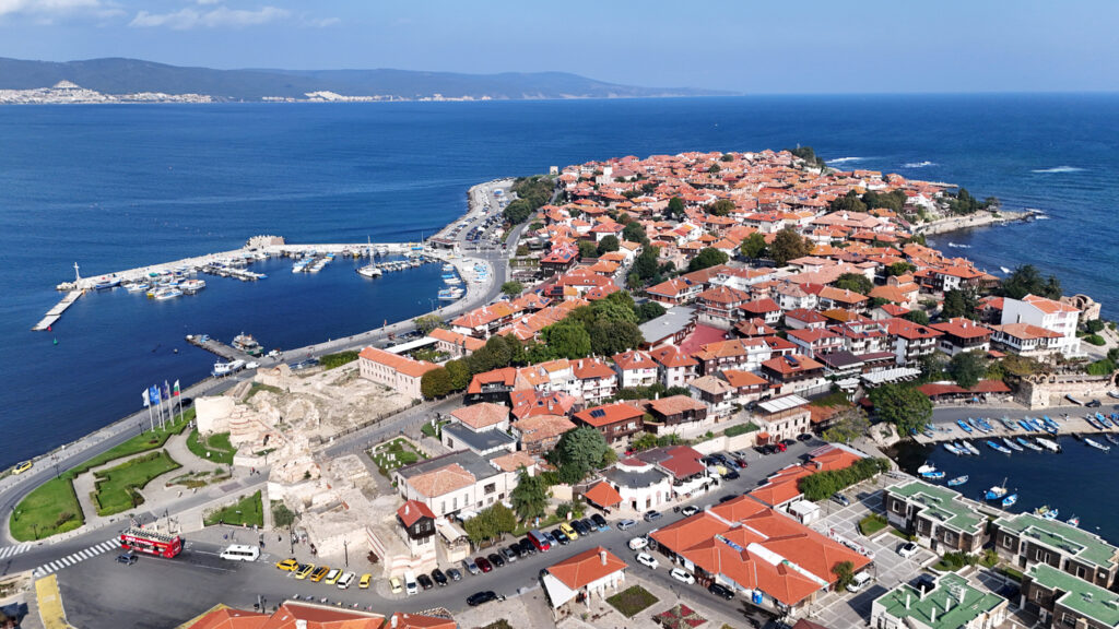 Luftaufnahme der Altstadt von Nessebar auf einer schmalen Halbinsel mit roten Ziegeldächern, zwei Häfen und Resten alter Stadtmauern im tiefblauen Meer.