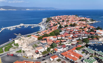 Luftaufnahme der Altstadt von Nessebar auf einer schmalen Halbinsel mit roten Ziegeldächern, zwei Häfen und Resten alter Stadtmauern im tiefblauen Meer.