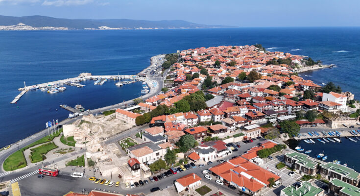 Luftaufnahme der Altstadt von Nessebar auf einer schmalen Halbinsel mit roten Ziegeldächern, zwei Häfen und Resten alter Stadtmauern im tiefblauen Meer.
