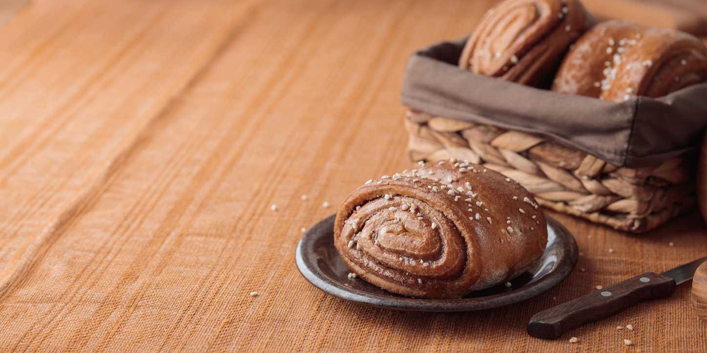 Einzelner Korvapuusti mit Hagelzucker auf Teller, dahinter Korb mit weiteren Zimtschnecken.