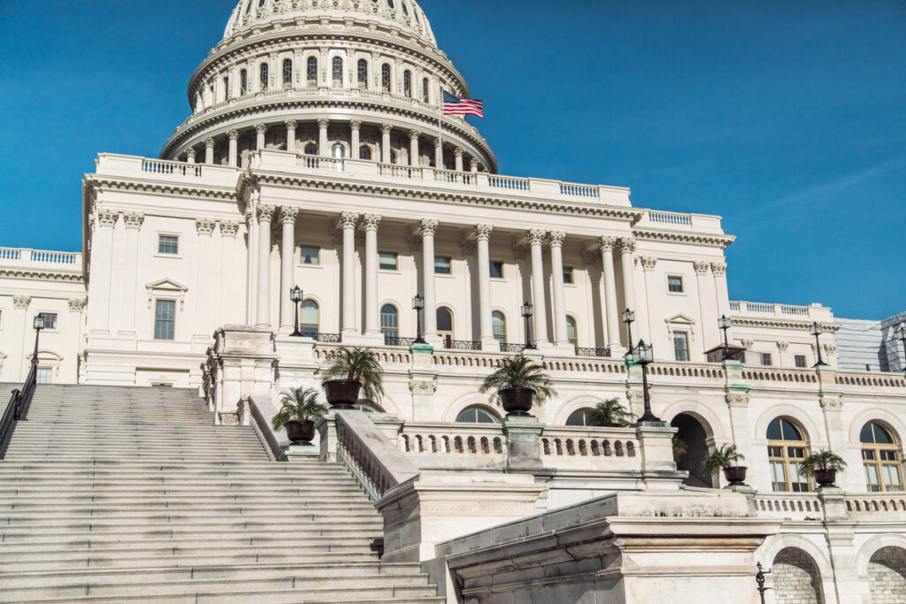 Nahaufnahme der Osttreppe des U.S. Capitols mit klassischer Säulenfassade und wehender US-Flagge vor blauem Himmel.