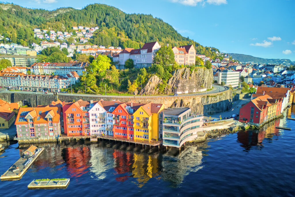 Luftaufnahme von Skuteviken mit farbigen Holzhäusern am Wasser, Felsstraße darüber und bewaldetem Hügel im Hintergrund.