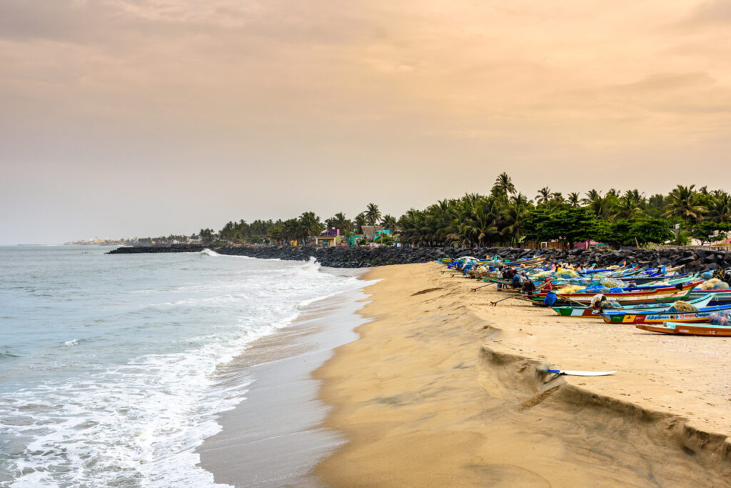Strand von Pondicherry in Indien mit bunten Fischerbooten, Palmen, Felswellenbrecher und aufgewühlter See im warmen Abendlicht.