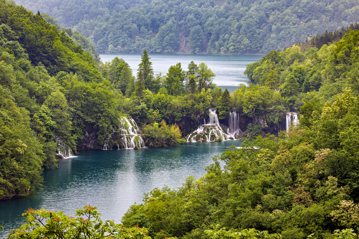 Mehrere Wasserfälle stürzen in einen türkisblauen See, umgeben von dichtem Wald.